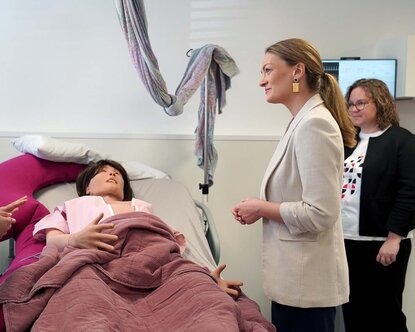 Gesundheitsministerin Judith Gerlach steht im Skills Lab vor einem Bett mit einer lebensgroßen Puppe, die eine schwangere Frau verkörpert, und spricht mit Professorin Hemma Pfeifenberger 
