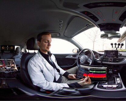 Forscher in weißem Kittel sitzt in einem Auto und zeigt etwas mit einem Stift auf einem Tablet.