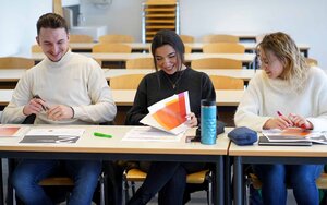 Ein Student und zwei Studentinnen sitzen nebeneinander an einem Tisch und sehen sich gemeinsam die Workshopmaterialien an. 