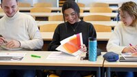Ein Student und zwei Studentinnen sitzen nebeneinander an einem Tisch und sehen sich gemeinsam die Workshopmaterialien an. 