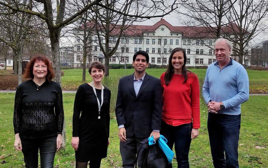 Prof. Dr. Renate Link, Jessica Albert, César Pancorvo Rosazza, Bettina Arnold und Prof. Dr. Holger Paschedag stehen nebeneinander im Freien vor der grünen Campuswiese, einigen unbelaubten Bäumen und einem weißen Hochschulgebäude mit rotem Dach im Hintergrund