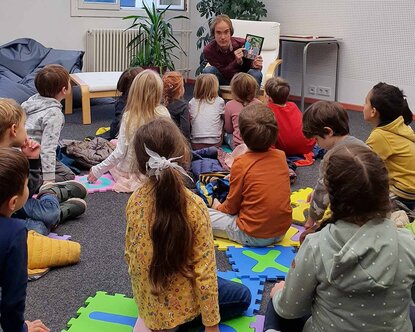 Ein Mann sitzt in einem Sessel und erzählt einer Gruppe von Kindern, die am Boden sitzen, eine Geschichte