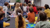 Ein Mann sitzt in einem Sessel und erzählt einer Gruppe von Kindern, die am Boden sitzen, eine Geschichte