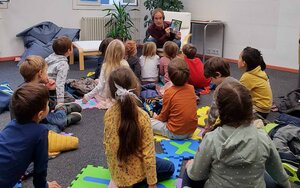 Ein Mann sitzt in einem Sessel und erzählt einer Gruppe von Kindern, die am Boden sitzen, eine Geschichte