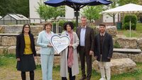 Frau mit herzförmigem Schild mit Spendensumme steht zwischen zwei Frauen und zwei Männern vor geöffnetem blauen Solarschirm