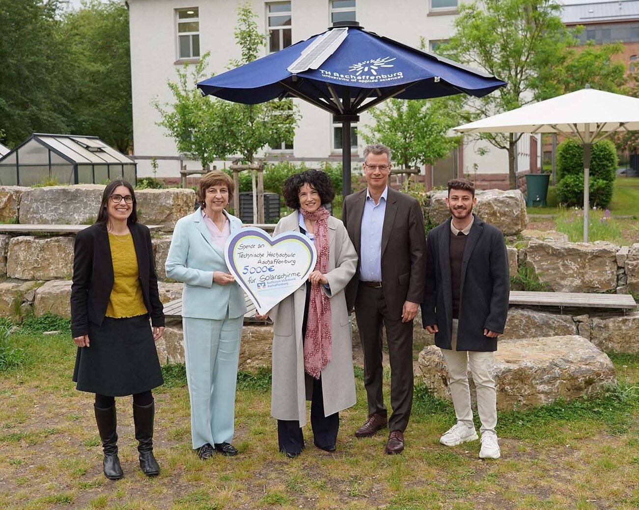 Frau mit herzförmigem Schild mit Spendensumme steht zwischen zwei Frauen und zwei Männern vor geöffnetem blauen Solarschirm