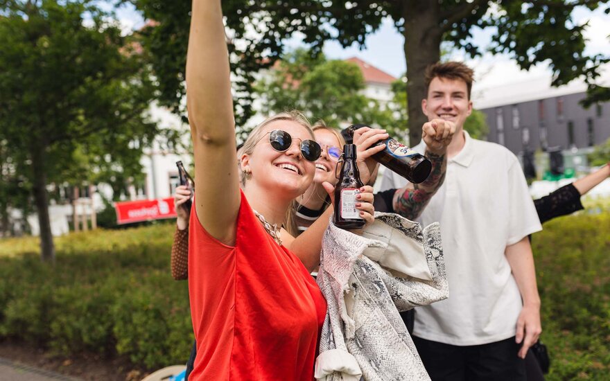 Drei Studierende feiern mit Bierflaschen auf dem Campus