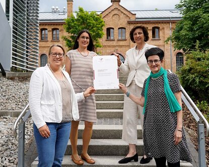 Vier Frauen stehen lächelnd auf einer Treppe vor einem Gebäude und halten ein Blatt Papier fest