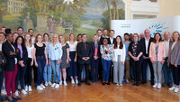 Eine Gruppe Studierenden sowie Professorinnen und Professoren in einem großen Saal der Hochschule vor einem Wandgemälde 