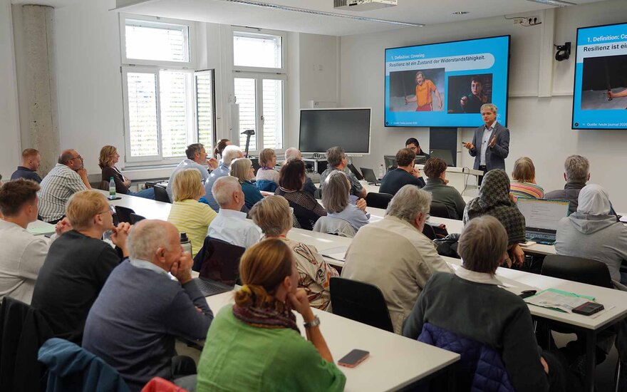 Publikum der Ringvorlesung von hinten sitzend an Tischen im Hörsaal mit Redner und Screens im Hintergrund