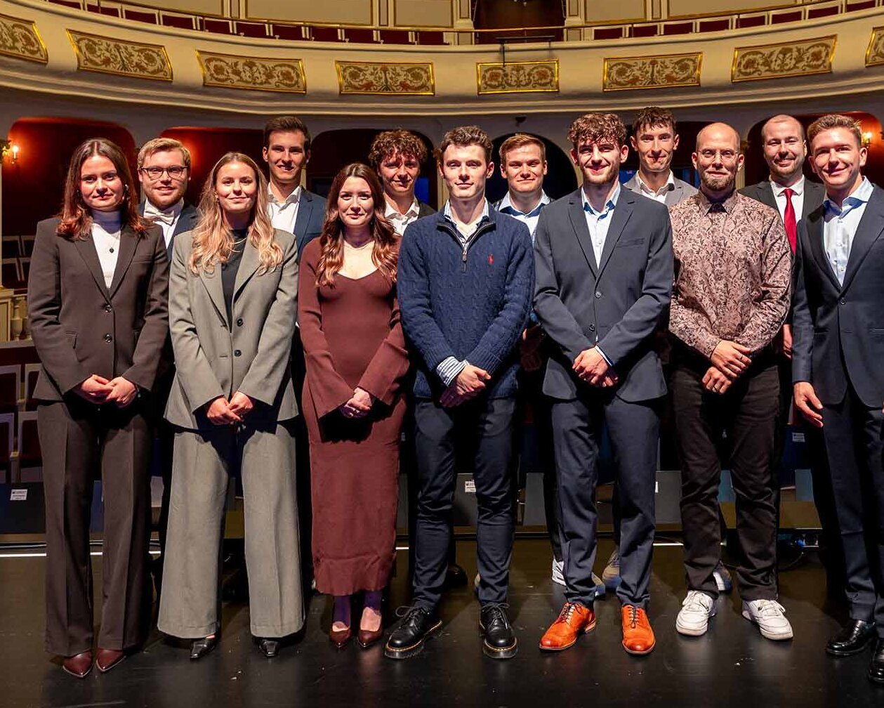 Gruppenbild mit jungen Männern und Frauen in Abendgarderobe auf der Bühne vor Zuschauerrängen im Theater