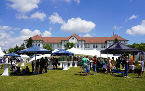 Gebäude der Hochschule im Hintergrund und Menschen unter Sonnenschirmen an Ständen im Vordergrund