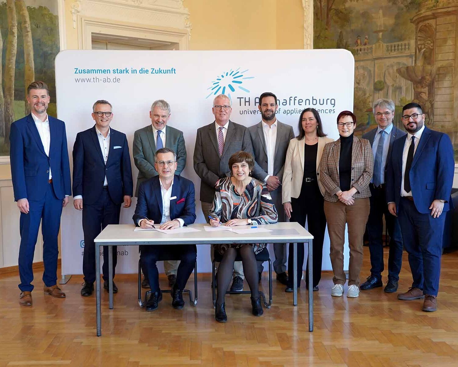 Zwei sitzende Personen, die Vertrag unterzeichnen und neun Personen stehen dahinter