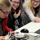 Schüler*innen sitzen an einem Schreibtisch und beschäftigen sich mit einem kleinen Roboter. 