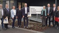 Gruppenbild mit 9 Personen vor dem Schild und dem Gebäude der Fakultät Gesundheitswissenschaften