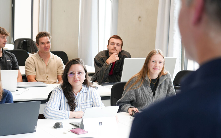 Mehrere Studierende sitzen in einem Seminarraum an Tischen mit Laptops und blicken in Richtung einer präsentierenden Person im Vordergrund. 