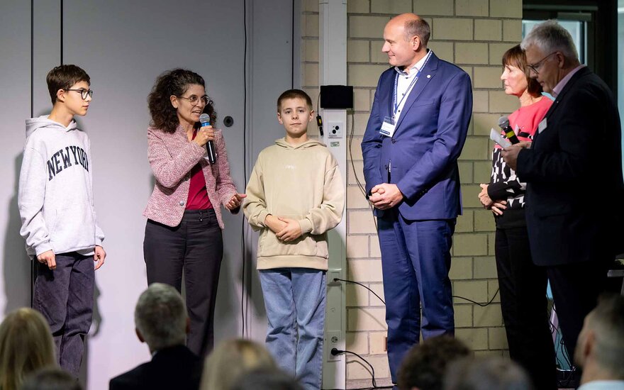 Schulleiterin spricht mit Mikrophon neben zwei Schülern, Mann mit blauem Anzug und einer weiteren Frau und einem Mann