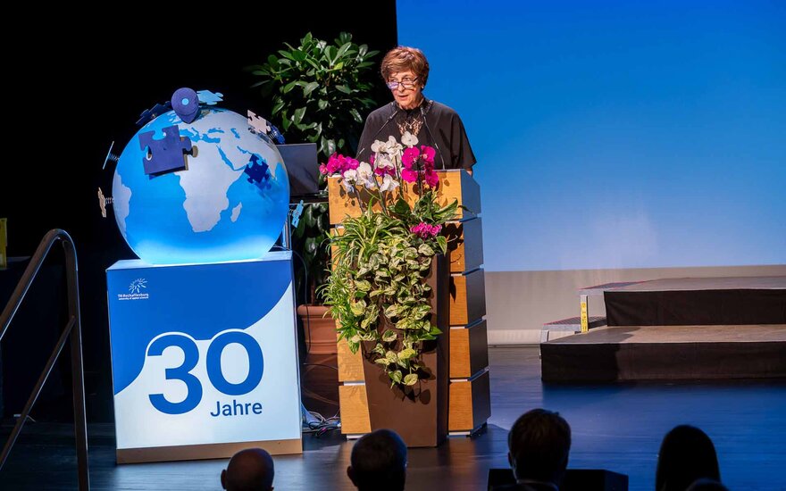 Dunkelhaarige Frau am Rednerpult mit Blumendeko und Globus mit 30-Jahre-Logo daneben