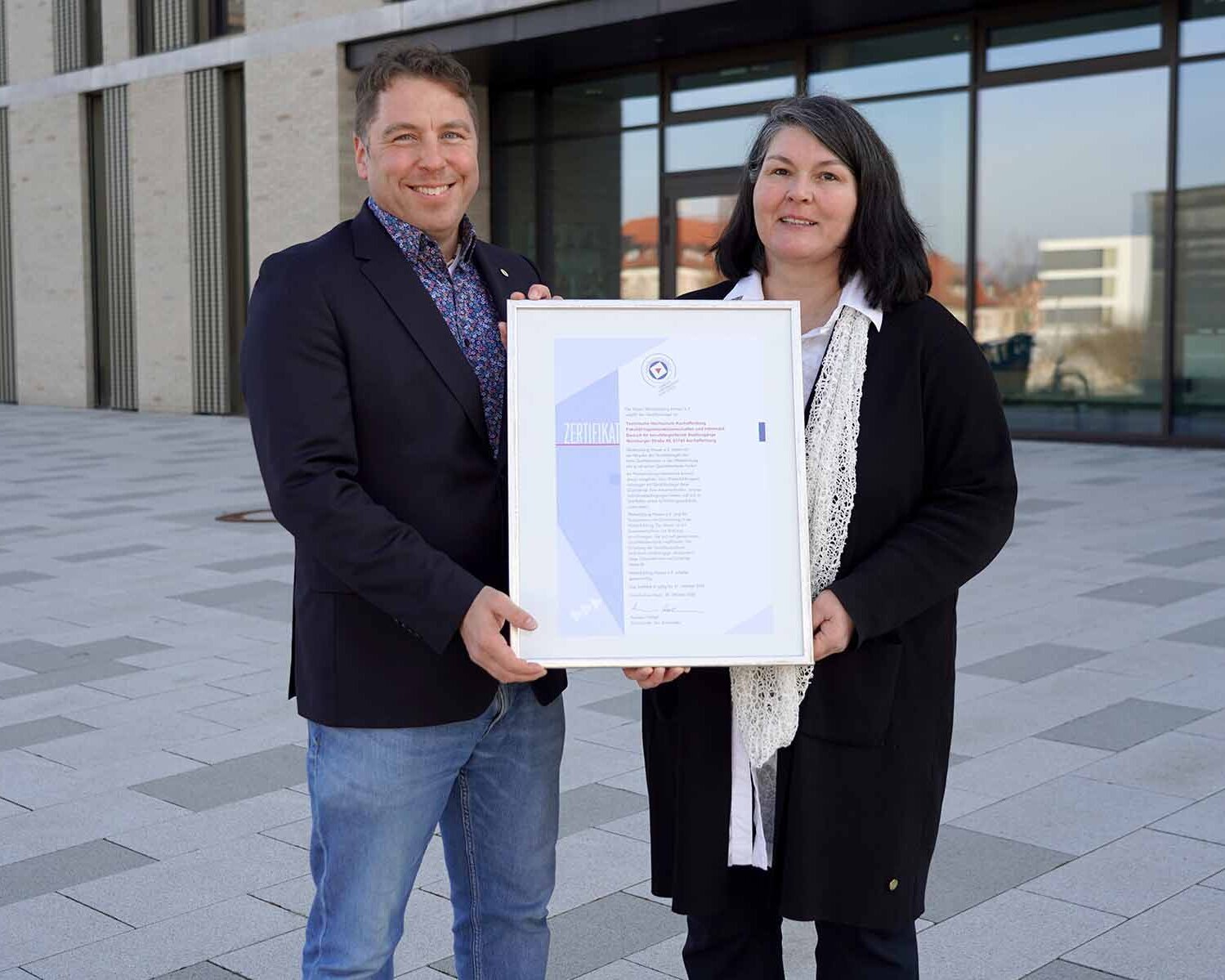 Ein Mann und eine Frau stehen vor einem TH-Gebäude mit der Urkunde in der Hand