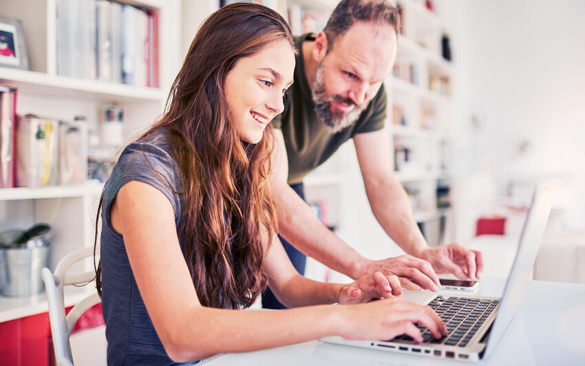 Eine junge lachende Frau sitzt an einem Laptop und ein älterer Mann steht hinter ihr und lacht auf den Bildschirm.