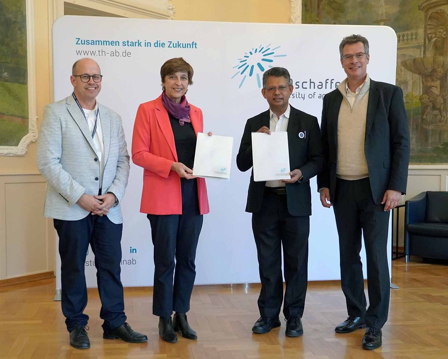 Frau mit rotem Blazer und Mann im schwarzen Anzug stehen mit Urkunden zwischen zwei Männern vor TH-Logowand