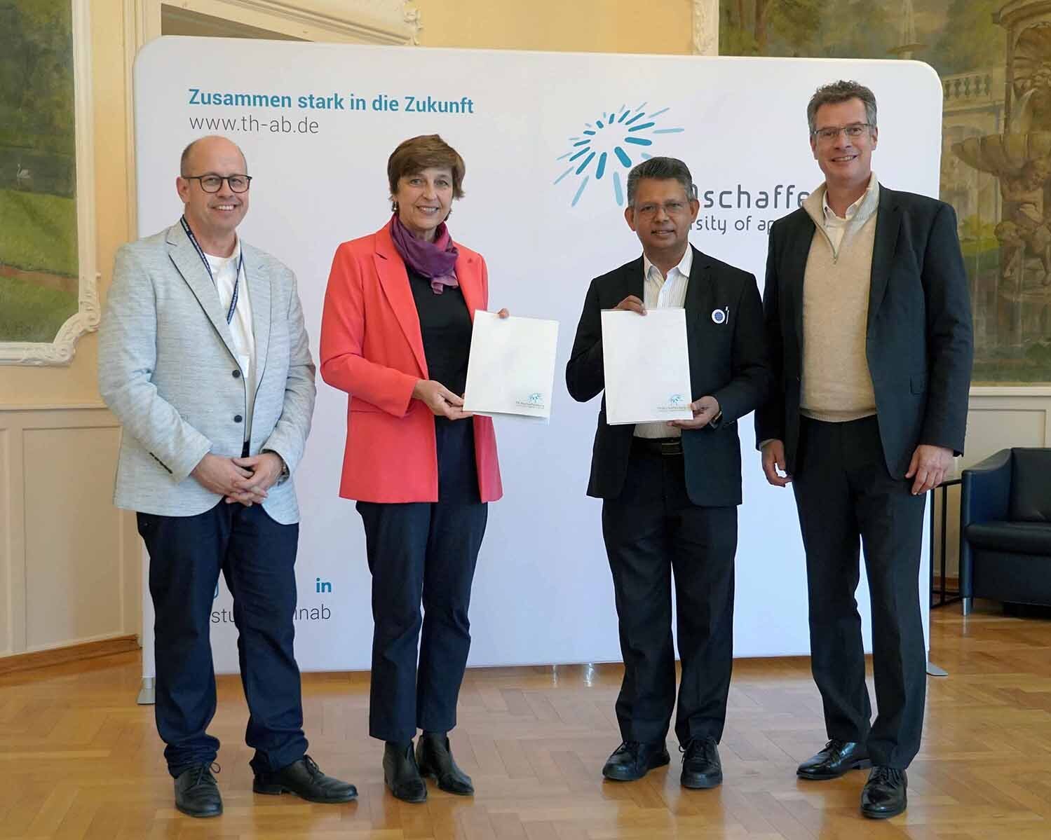 Frau mit rotem Blazer und Mann im schwarzen Anzug stehen mit Urkunden zwischen zwei Männern vor TH-Logowand