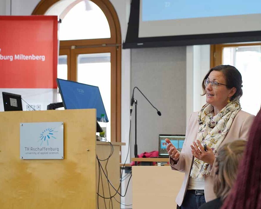 Professorin Bertels spricht am Rednerpult mit TH-Logo in der Aula, rotes Sparkassen-Roll-up dahinter