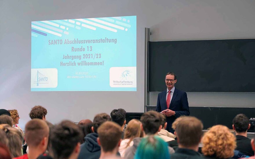 Professor Mewes steht vor einer Tafel und einer Projektion der blauen Begrüßungspräsentation und spricht zu den sitzenden Schülerinnen und Schülern.