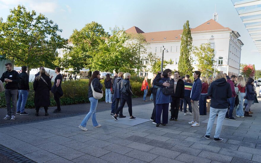 Gruppe von Personen stehen vor Bäumen und einem von der Sonne angestrahlten weißen Hochschulgebäude mit rotem Ziegeldach und unterhalten sich