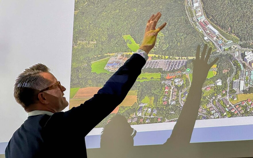 Ein Mann präsentiert vor einer projizierten Luftaufnahme eines Gebiets und zeigt mit ausgestrecktem Arm auf markierte Bereiche; auf der Leinwand sind Waldflächen, Gebäude und geplante Strukturen erkennbar.