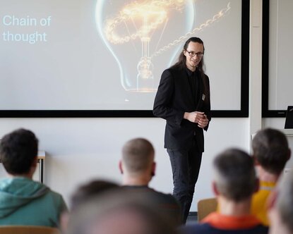 Ben Lenk-Ostendorf bei seinem Impulsvortrag mit Blick zum Publikum, im Hintergrund eine Glühbirne auf der Leinwand mit Überschrift Chain of thought