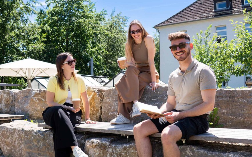 Drei Personen sitzen im Außenbereich auf gestuften Steinblöcken in einem Hochschulgarten und betrachten gemeinsam Inhalte in einem Notizbuch; im Hintergrund sind Bäume, ein Gewächshaus und ein Sonnenschirm zu sehen.