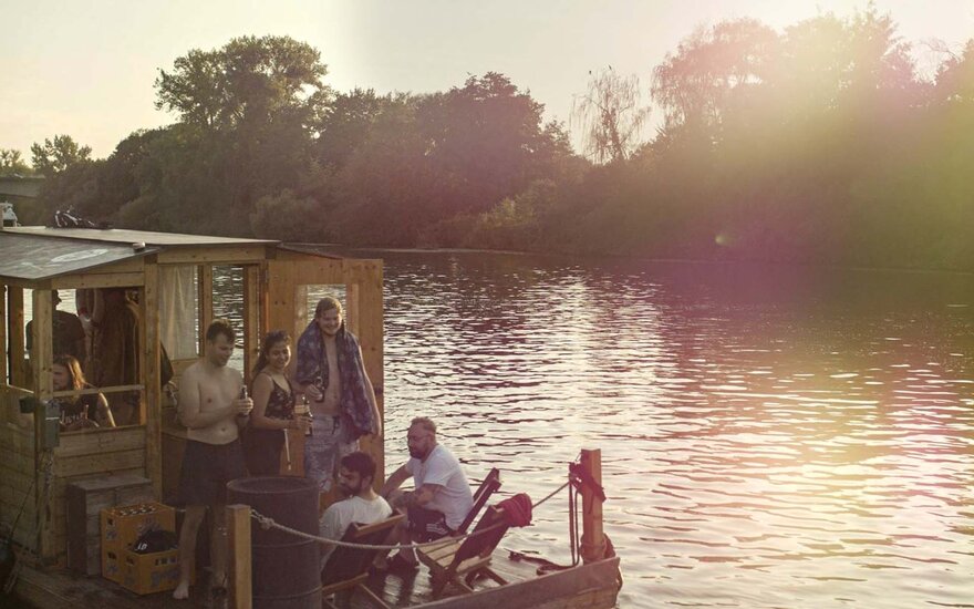 Sieben Studierende Gruppe auf hölzernem Hausboot am Steg auf dem Wasser bei Sonnenuntergang, gemütliches Beisammensein lachend und mit Flasche in der Hand