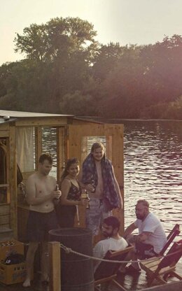 Sieben Studierende Gruppe auf hölzernem Hausboot am Steg auf dem Wasser bei Sonnenuntergang, gemütliches Beisammensein lachend und mit Flasche in der Hand