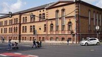 Zwei Personen mit Kinderwagen, zwei Kinder mit Rollern, ein Fahrradfahrer und zwei Autos an Ampelkreuzung vor braunem Backsteingebäude der TH