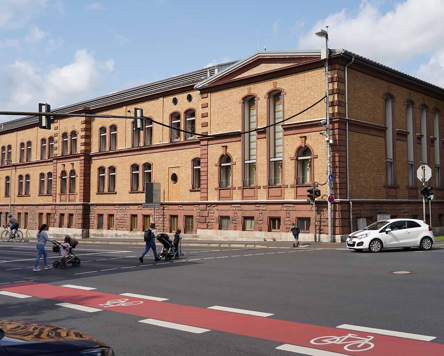 Zwei Personen mit Kinderwagen, zwei Kinder mit Rollern, ein Fahrradfahrer und zwei Autos an Ampelkreuzung vor braunem Backsteingebäude der TH