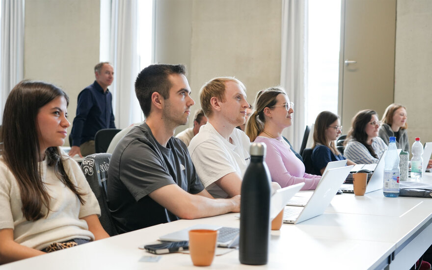 Mehrere Studierende sitzen an Tischen in einem hellen Vorlesungsraum und arbeiten mit Laptops, während sie aufmerksam in Richtung einer Präsentation blicken. 