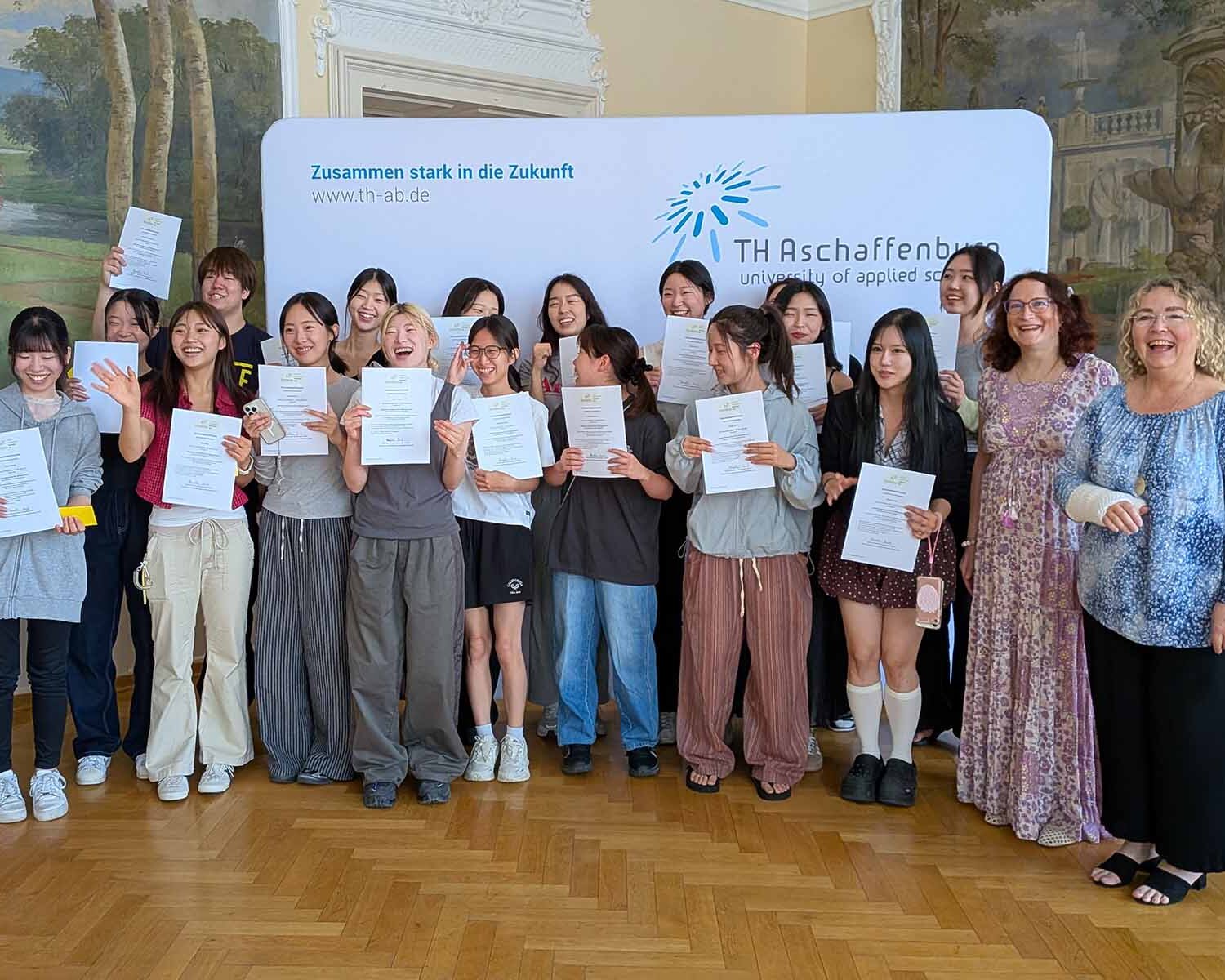 Gruppenfoto mit 20 Personen mit Zertifikaten im Hocksaal vor Logowand