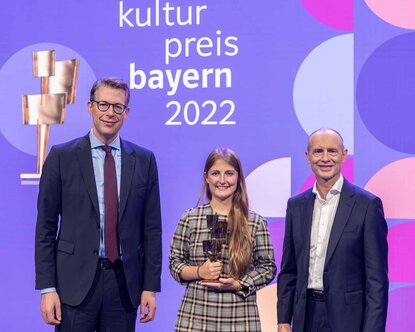 Nebeneinander vor einer violetten Wand mit der Aufschrift Kulturpreis Bayern 2022 steht ein Mann mit dunklem Anzug und Brille, eine junge Frau mit langen dunkelblonden Haaren und einer goldenen Statue in der Hand und ein Mann mit Glatze und ebenfalls dunklem Anzug