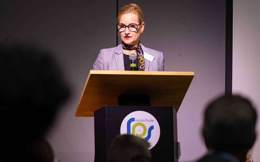 Frau mit hochsteckten Haaren und Brille spricht am Rednerpult