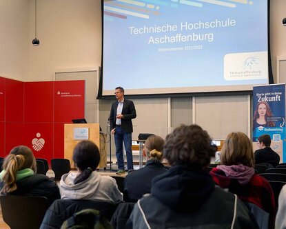 Ein Professor der Fakultät Wirtschaft und Recht steht in der Aula neben dem Rednerpult und spricht zu den sitzenden Schülerinnen und Schülern. Im Hintergrund ist auf einer blauen Leindwand der Schriftzug der TH Aschaffenburg zu sehen. Daneben steht einer rote Wand der Sparkasse.