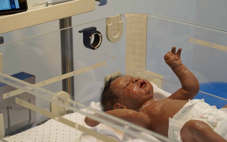 Eine dunkelhäutige Simulationspuppe in Größe eines Babys liegt in einem Bett mit einer Glasumrandung