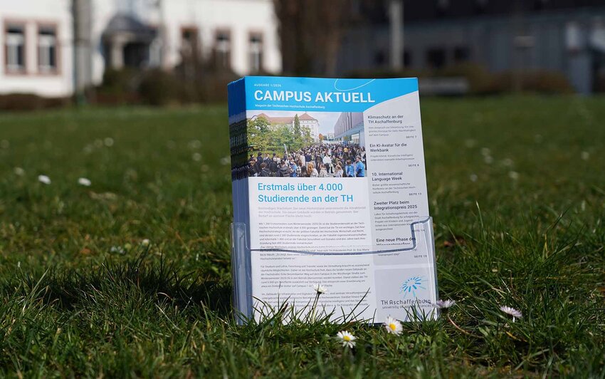 ochschulmagazine im Prospektständer auf Wiese mit Gänseblümchen vor Gebäuden der TH