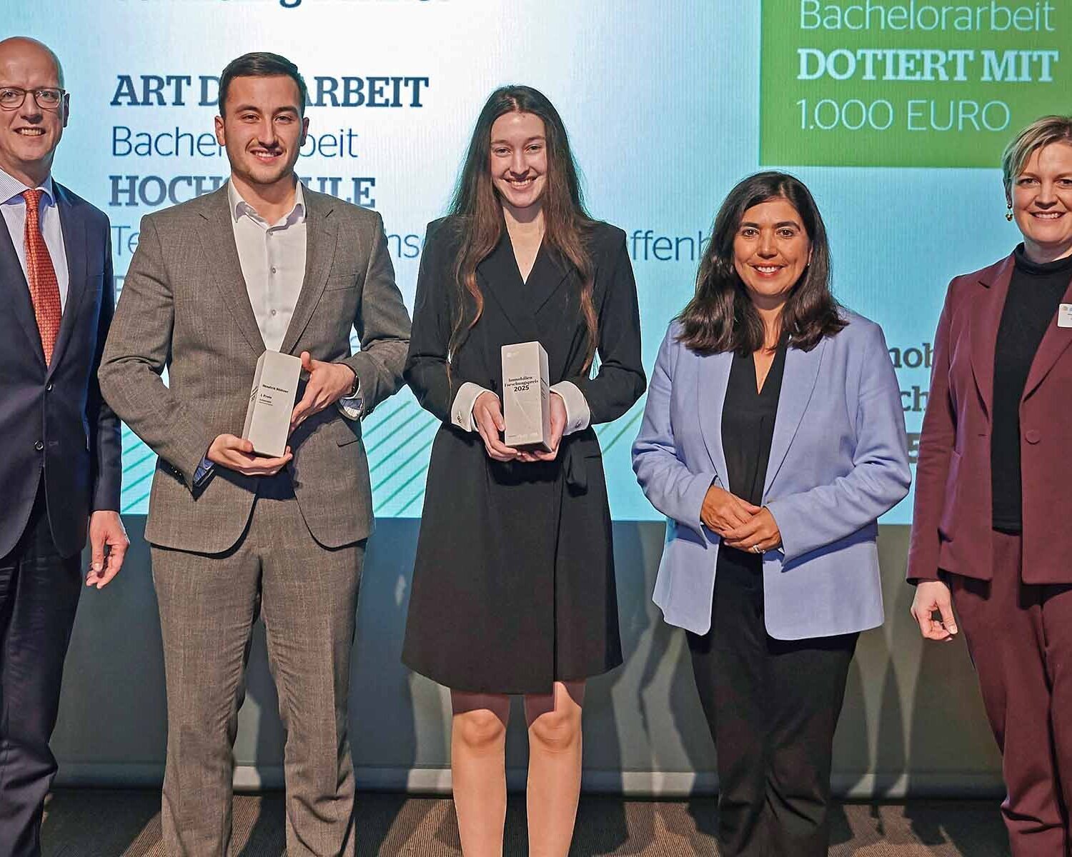 Fünf Menschen stehen vor einer Leinwand. Zwei von ihnen halten Preise in der Hand