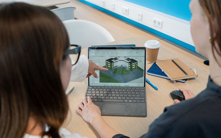 Zwei Studentinnen sitzen an einem Tisch und betrachten gemeinsam ein Tablet, auf dem eine 3D-Visualisierung eines Gebäudekomplexes mit Innenhof angezeigt wird; eine Person zeigt mit dem Finger auf das Modell, während auf dem Tisch ein Kaffeebecher, ein Notizbuch und ein Stift liegen.