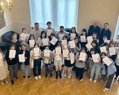 Gruppe der internationalen Kursteilnehmenden, die ihre Zertifikate hochhalten, im Hocksaal der Hochschule