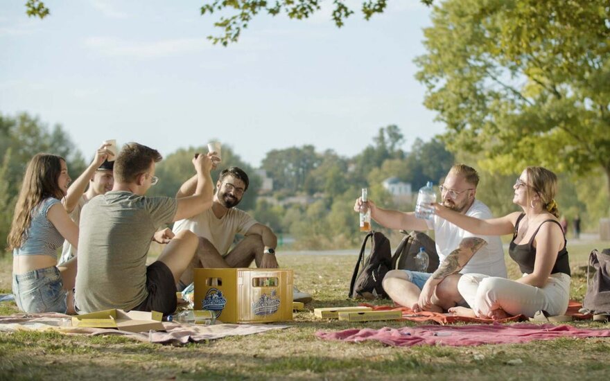 Sechs Studierende Gruppe sitzen auf Wiese bei Sonnenschein, heben die Getränke zum Toast, trinken Bier und Wasser