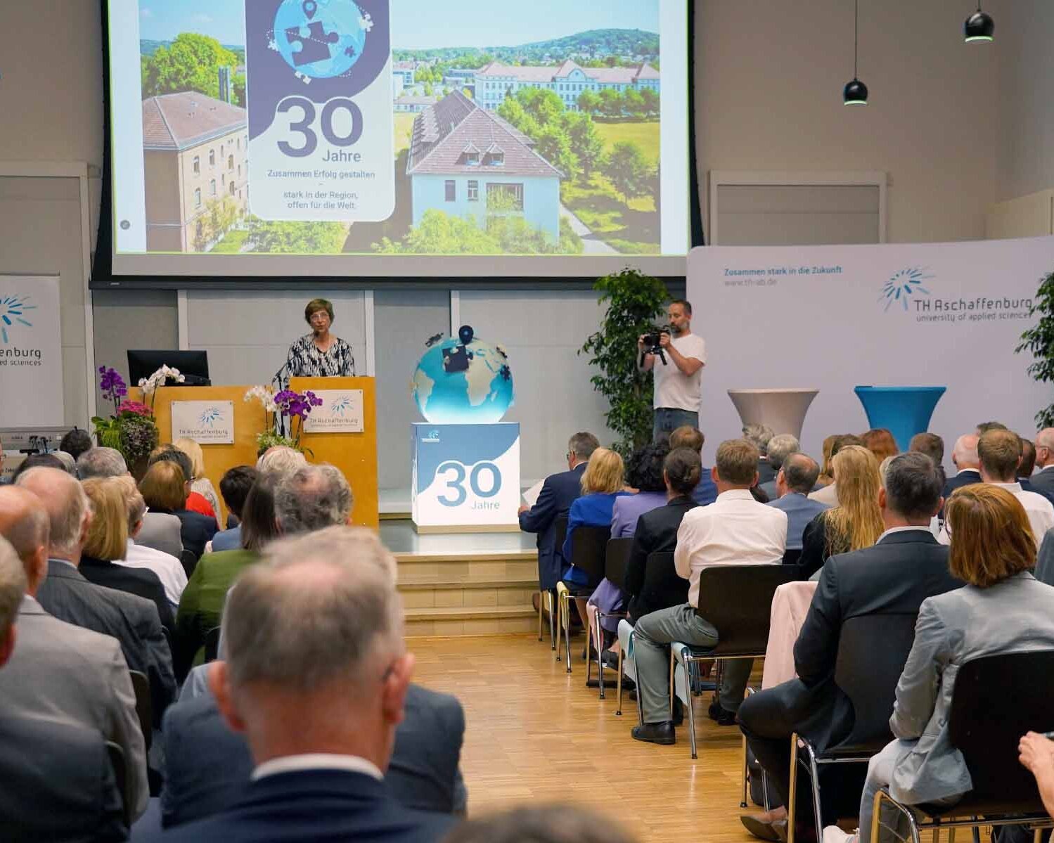 Präsidentin mit Keyvisual im Hintergrund und als Plastik auf dem Boden neben Rednerpult vor Publikum in der Aula