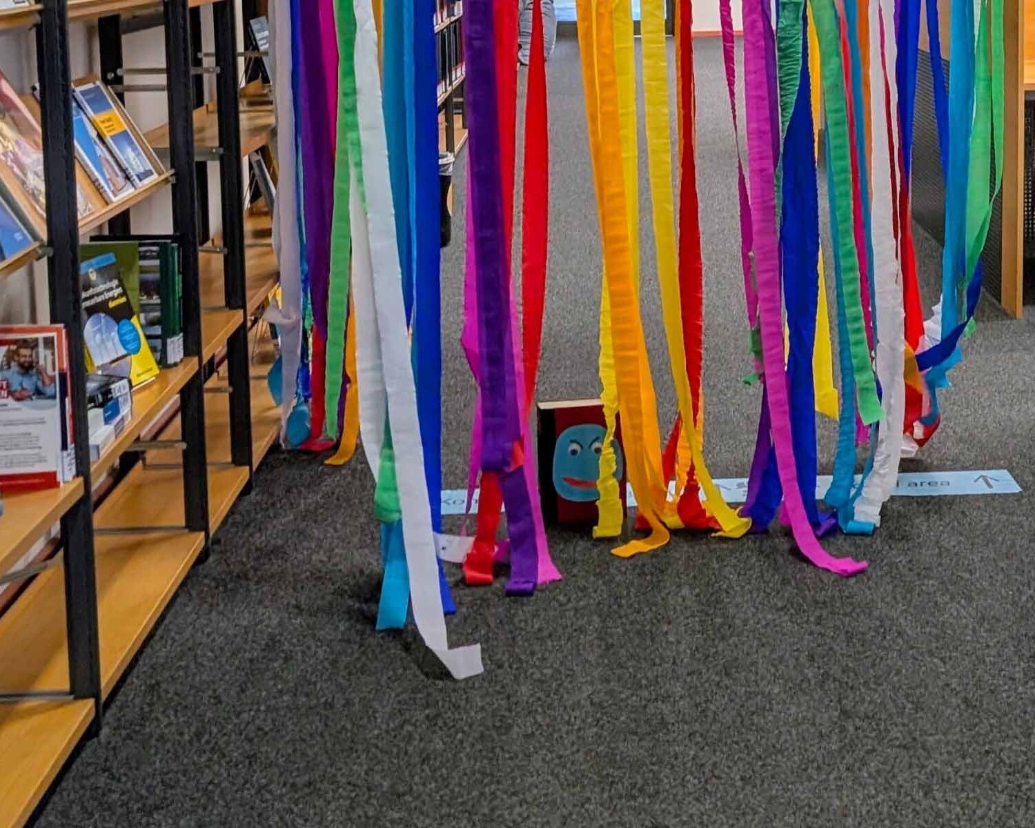 ein gebastelter Bücherwurm mit vielen bunten Papierstreifen auf dem Boden vor dem Bücherregal