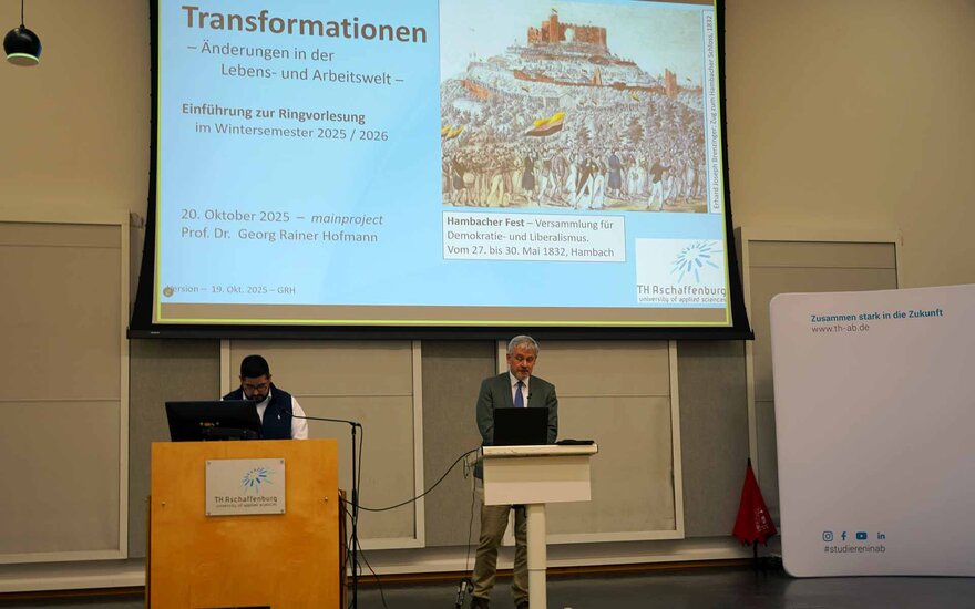 Ein Mann im Anzug steht hinter einem Rednerpult. Im Hintergrund Präsentation mit Titel „Transformationen“. 